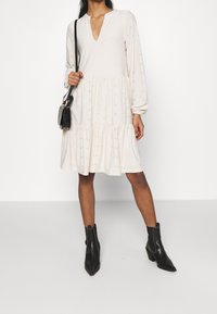 Robe crème avec un décolleté en V, des manches longues et un ourlet en couches. Présente des petits trous décoratifs. Associée à des bottines noires et un sac à main.