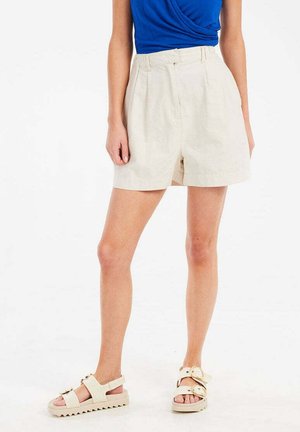 Beige linnen shorts met een hoge taille en geplooide voorkant. Gepaard met een blauwe top en lichtgekleurde sandalen met banden en een chunky zool.