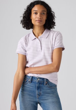 Junge Frau mit lockigem schwarzen Haar, die ein hellviolettes gestreiftes Poloshirt und blaue Jeans trägt, steht mit verschränktem Arm.