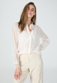 Weiße Bluse mit auffälliger Spitzenverzierung an den Ärmeln und der Vorderseite, aus leichtem Material, kombiniert mit beigen Hosen.