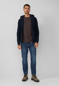 Sudadera con cremallera de rayas azul marino sobre una camiseta de rayas marrón, combinada con vaqueros azules y botas marrones con cordones. Un atuendo simple y casual.