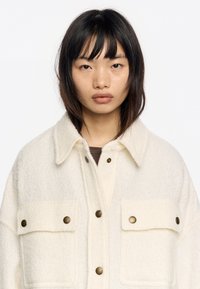 Chaqueta de lana color crema con diseño de cuello, que cuenta con dos bolsillos en el pecho, botones de presión de latón y un acabado de tejido texturizado.