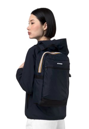 Mladá žena v tmavej bunde a bielych nohaviciach nesie námornícky modrý batoh so žltými a bielymi pruhmi na popruhoch.