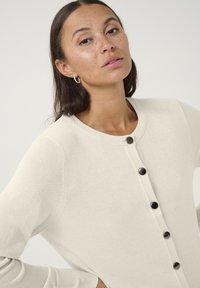 Kremni kardigan iz mehkega blaga, z okroglim izrezom in črnimi gumbi spredaj. Dolgi rokavi in rebrasti manšeti.