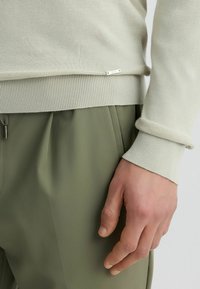 Svijetlosivi pulover s rebrastim rubom i manžetama, napravljen od mekanog pletenog materijala. Ima malu metalnu oznaku blizu donjeg ruba. Maslinasto zeleni hlače.