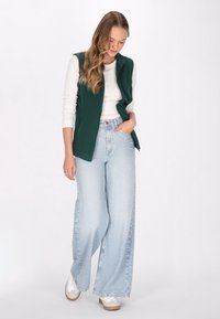Grön ärmlös väst över en vit långärmad skjorta, i kombination med ljusblå vida jeans och vita sneakers. Enkelt mönster och avslappnad stil.