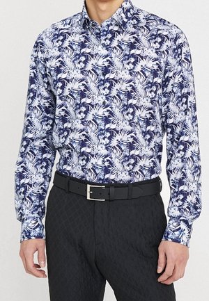 Man draagt een lange mouw blouse met knopen met een blauw-wit bloemenpatroon, zwarte riem en zwarte gestructureerde broek.