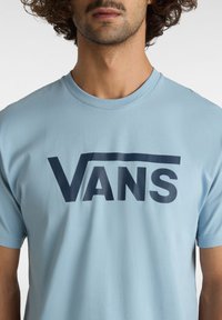Ljusblå T-shirt med korta ärmar som har en djärv marinblå "VANS"-logotyp över bröstet. Mjuk bomullstyg med rund halsringning.