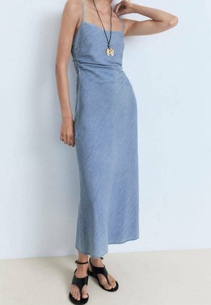 Femme portant une robe longue sans manches en denim bleu clair avec des fines bretelles et des sandales noires à bouts ouverts, debout contre un mur blanc.