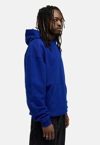 Blaue Kapuzenjacke aus weichem Stoff, mit einer Fronttasche, großer Kapuze und gerippten Bündchen. Getragen mit dunklen Jeans.