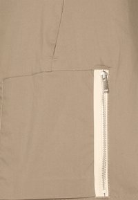 Tissu de veste beige avec une texture lisse, présentant un détail de poche zippée blanche bien visible sur le côté. Design minimaliste avec des lignes épurées.