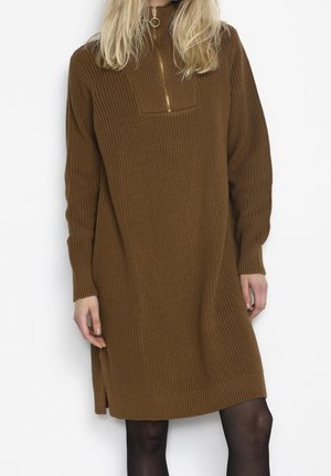 Kvinde iført en brun ribstrikket sweaterkjole med en forgyldt lynlås foran og lange ærmer, kombineret med sorte gennemsigtige strømpebukser.