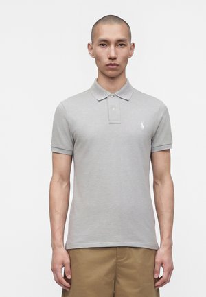 Mand med kort hår iført en lysegrå poloshirt og brune bukser, står foran en ensfarvet hvid baggrund.