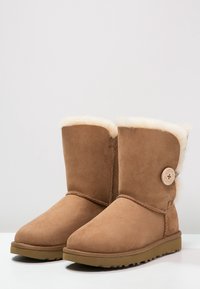 Par de botas de ante marrón hasta la mitad de la pantorrilla con forro de felpa en color crema, detalle de botón de madera redondo y suelas de goma planas.