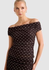 Femme blonde portant un haut noir à épaules dénudées avec des pois blancs et des boucles d'oreilles hoop en or, regardant sur le côté sur un fond uni.