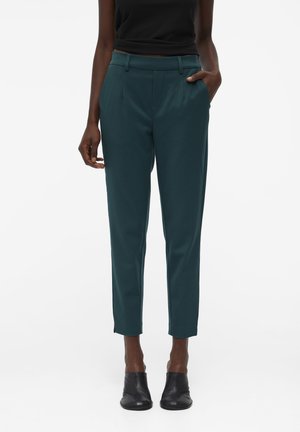 Vrouw draagt donkerteal taps toelopende broek met zijzakken en zwarte enkellaarsjes, staand tegen een effen witte achtergrond.