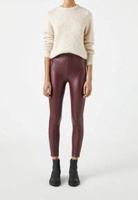 Kvinna i krämfärgad, fluffig tröja, högmidjade bordeaux läderbyxor och svarta ankelstövlar, stående framför vit bakgrund.