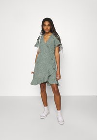 Vestido verde envolvente com bolinhas brancas, mangas curtas com babados e uma barra rodada. Combinado com sapatilhas brancas até ao tornozelo.