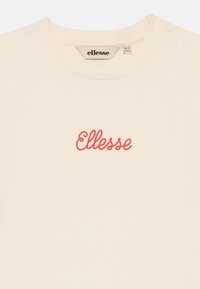 Krémové bavlněné tričko s krátkým rukávem a kulatým výstřihem. Na hrudi se nachází červené logo "Ellesse" napsané kurzívou.