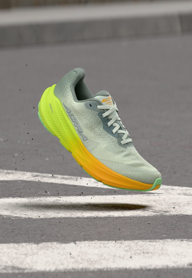 Αθλητικό παπούτσι ανοιχτού γκρι με νέον κίτρινο, στον αέρα πάνω από άσφαλτο με λευκές γραμμές, με εμφανές το πέλμα και το υφασμάτινο επάνω μέρος με υφή.
