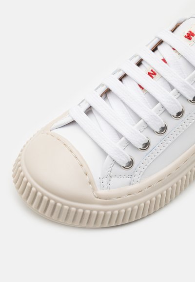 Sneaker bianco con una punta rotonda in gomma beige, lacci bianchi, occhielli argentati e una suola in gomma texturizzata. Presenta un sottile dettaglio di cucitura blu.