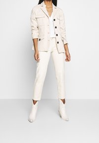 Chaqueta beige de botones con múltiples bolsillos, combinada con pantalones ajustados de color claro y botas blancas hasta el tobillo con un diseño cortado.