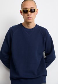 Marineblauwe textuurtrui met raglanmouwen en ronde hals, gecombineerd met geel getinte zonnebril. Model staat met handen gevouwen.