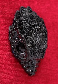 Czarna, dekoracyjna broszka w kształcie ręki, z błyszczącymi teksturami i osadzonymi czarnymi kamieniami o różnej wielkości na czerwonym, teksturowanym tle.