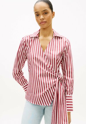 Kobieta ubrana w czerwoną i białą bluzkę z pionowymi paskami na długim rękawie, zawiązaną w talii, oraz jasnoniebieskie dżinsy, stojąca na jednolitym tle.