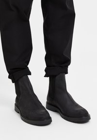 Černé semišové chelsea boty s elastickými bočními panely a gumovou podrážkou. Kalhoty jsou černé, mírně ohrnuté na nohavicích, což odhaluje boty.