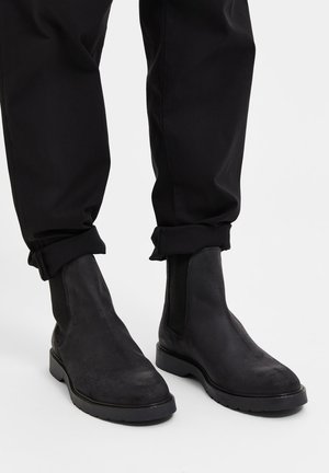 Stivaletti Chelsea in suede neri con pannelli elastici laterali e suola in gomma. I pantaloni sono neri, leggermente arrotolati alle estremità, mostrando gli stivaletti.
