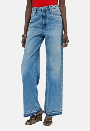 Personne portant un jean bleu taille haute à jambes larges, un haut rouge, des sandales compensées à motif léopard, un bracelet en or et des bagues en argent sur fond clair.