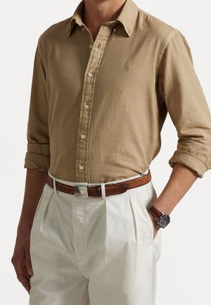 Man wearing beige button-up shirt with rolled sleeves, white pleated trousers, brown belt, and brown wristwatch with black dial.