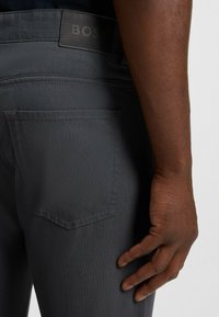 Grå tygbyxor med en strukturerad finish. Har en bakficka och en mörk logotagg. Närbild av en hand som vilar på höften.