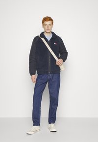 Giacca in pile blu navy con zip, maniche a contrasto e patch con logo. Indossata sopra una camicia azzurro chiaro e abbinata a jeans scuri e sneaker.
