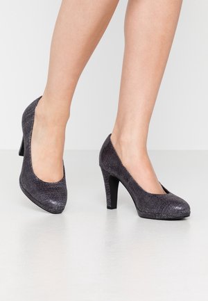 Kvinde iført mørkegrå strukturerede højhælede sko, der står på et lyst gulv mod en ensfarvet hvid baggrund.