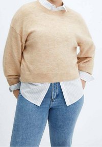 Beige gestrickter Pullover mit rundem Halsausschnitt, kombiniert mit einem weiß gestreiften Hemd mit Kragen darunter. Getragen mit hellblauen High-Waist-Jeans.