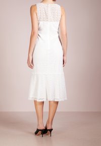 Vit klänning med texturerad spetsliv och utsvängd kjol. Har en bakre dragkedja och nederkant som slutar under knäet. Bärs med svarta högklackade skor.