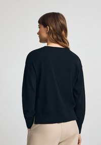 Suéter negro de punto con corte relajado, cuello redondo, hombros caídos y dobladillo acanalado, combinado con pantalones de color claro.
