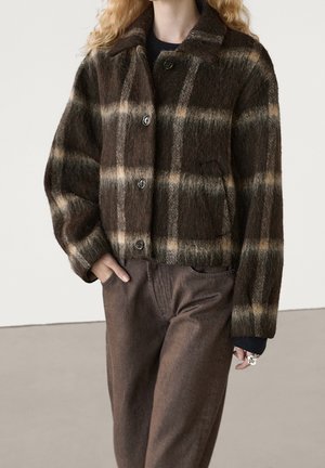 Personne portant une veste en laine à carreaux marron et beige boutonnée avec un pantalon marron assorti, une main dans la poche et des bagues aux doigts.