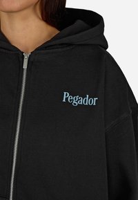 Čierna mikina s kapucňou na zips vyrobená z bavlny s uvoľneným strihom. Na hrudi sa nachádza svetlomodré logo "Pegador" a má stahovaciu kapucňu.
