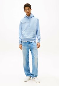 Felpa azzurra chiara con tasca frontale, cordino regolabile e una striscia nera orizzontale. Abbinata a jeans azzurri larghi e sbiaditi e sneakers bianche.