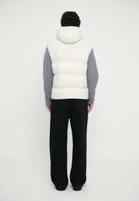 White puffer vest with a hood and grey wool sleeves, featuring a tonal "EA7" logo on the back. Worn with black pants.