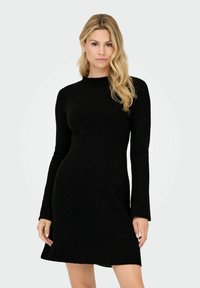 Frau mit langen blonden Haaren, die ein figurbetontes schwarzes Rippstrickkleid mit langen Ärmeln trägt, steht vor einem schlichten hellen Hintergrund.