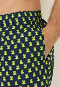 Costumi da bagno blu navy con un motivo di rane verdi, alcune delle quali indossano accessori. Vita elasticizzata con tasche; tessuto liscio e leggero.