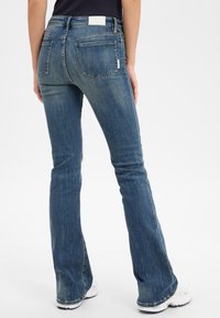 Vaqueros de corte acampanado en denim azul medio con un acabado deslavado, que presentan bolsillos traseros y una etiqueta blanca en la cinturilla.