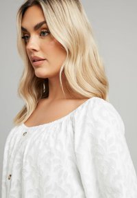 White blouse with textured floral pattern, off-the-shoulder neckline, gathered elastic trim, and button accents. Soft fabric and flowing fit.
