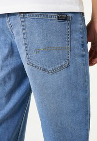 Jeans de mezclilla azul claro con un bolsillo trasero que presenta una línea de costura distintiva. La tela tiene una textura suave y un ligero desgaste.
