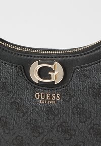 Black textured handbag with gold zipper and hardware. Features prominent logo, "GUESS EST 1981," embossed in gold. Elegant design and finish.