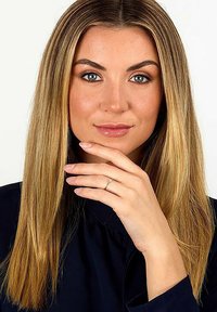 Eine Person mit langen, glatten, hellbraunen Haaren, die ein schwarzes Oberteil trägt, posiert mit einer Hand nahe ihrem Gesicht und einem schlichten Silberring an einer Hand.